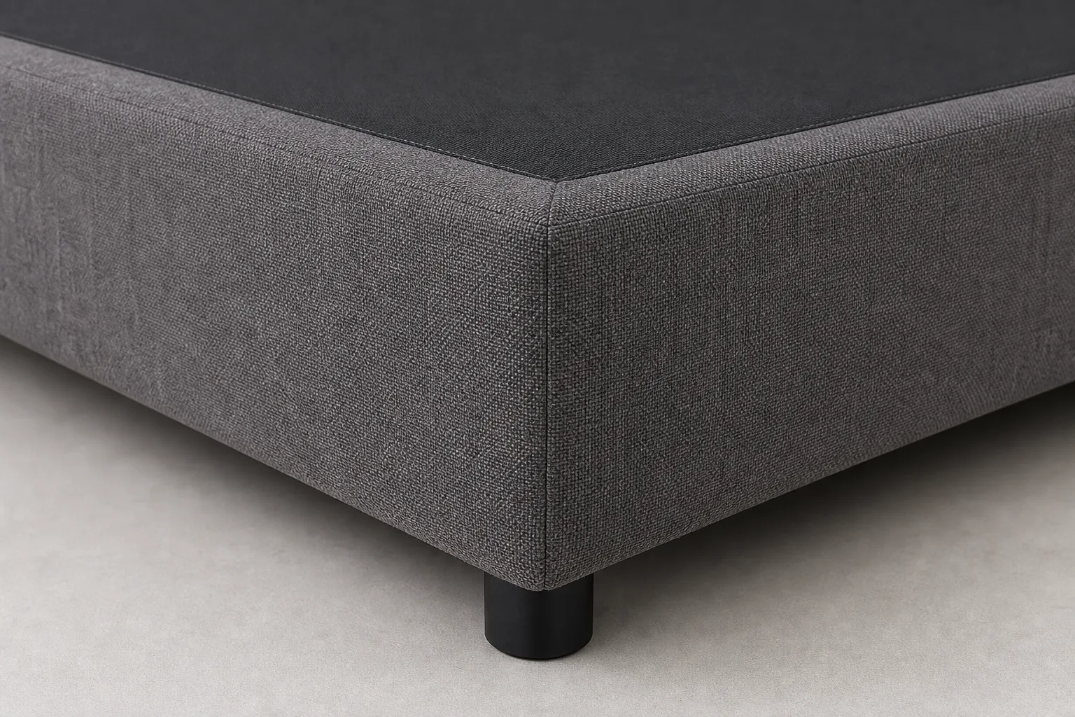 Close-up of grey upholstered hotel mattress base corner with woven fabric seam and black plinth leg